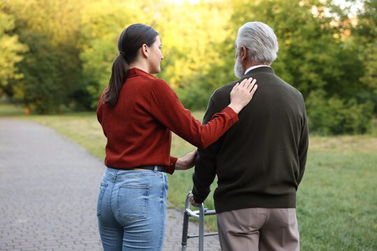 Caregiver supporting elderly man who using walking frame outdoors, back view