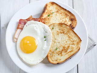 A plate contains a fried egg, some pieces of bacon, and two slices of toasted bread, making for a...
