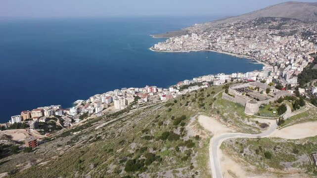 Scenic aerial view of Sarande cityscape on shores of gulf of Ionian Sea and ancient Lekuresi Castle on hill above city on sunny spring day, Albania 