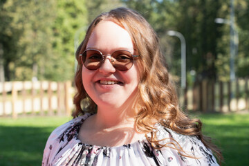 Smiling young woman with long curly hair and sunglasses standing outdoors on a sunny day, looking at the camera.