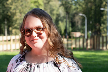 Smiling young woman with long curly hair and sunglasses standing outdoors on a sunny day, looking...