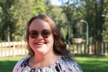 Smiling young woman with long curly hair and sunglasses standing outdoors on a sunny day, looking...