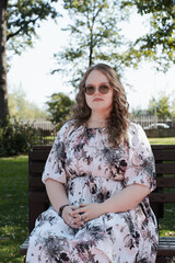 Young woman in a floral dress and sunglasses sitting on a park bench on a sunny day, looking at the...