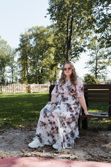 Young woman in a floral dress and sunglasses sitting on a park bench on a sunny day, looking at the...
