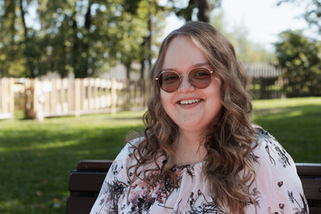 Young woman in a floral dress and sunglasses sitting on a park bench on a sunny day, looking at the...
