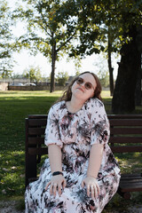 Young woman in a floral dress and sunglasses sitting on a park bench on a sunny day, looking at the...