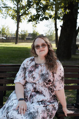 Young woman in a floral dress and sunglasses sitting on a park bench on a sunny day, looking at the...