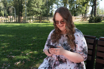 Young woman with long curly hair using a smartphone outdoors in a park on a sunny day, smiling and...