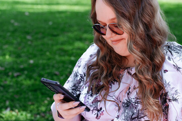 Young woman with long curly hair using a smartphone outdoors in a park on a sunny day, smiling and...