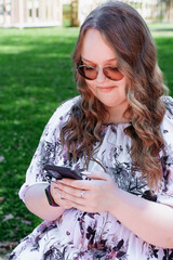 Young woman with long curly hair using a smartphone outdoors in a park on a sunny day, smiling and...