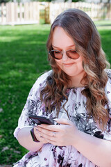 Young woman with long curly hair using a smartphone outdoors in a park on a sunny day, smiling and...