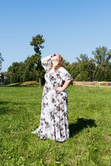 A young woman in a floral dress and sunglasses is posing joyfully on a grassy field in the park on a sunny day