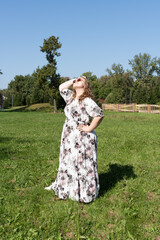 A young woman in a floral dress and sunglasses is posing joyfully on a grassy field in the park on...
