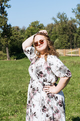 A young woman in a floral dress and sunglasses is posing joyfully on a grassy field in the park on a sunny day
