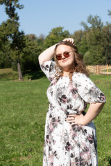 A young woman in a floral dress and sunglasses is posing joyfully on a grassy field in the park on a sunny day