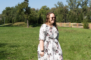 A young woman in a floral dress and sunglasses is posing joyfully on a grassy field in the park on a sunny day