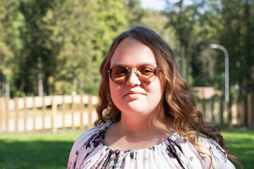 Smiling young woman with long curly hair and sunglasses standing in the park on a sunny day,...