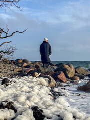 Individual standing on rocky shoreline, gazing at ocean waves, wearing a long black coat and white...