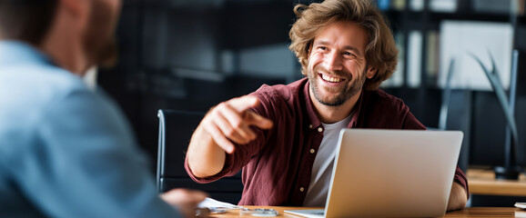 Cheerful Office Meeting: An enthusiastic man gestures expansively in a meeting while a laptop sits ready for action on the table, embodying collaboration in the modern workspace.