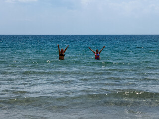 Two women enjoying a sunny day in the ocean, splashing water joyfully while standing in shallow...