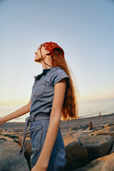 lifestyle woman in modern boho-western denim outfit with red headscarf stands by rocky beach at...