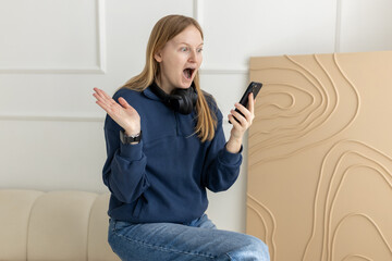 A young blonde woman employee with a happy successful expression holding her phone and sitting on coach at home