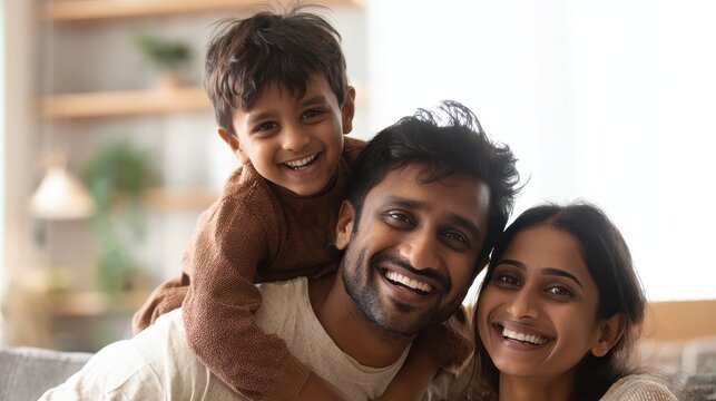 Cheerful indian son sitting on father shoulder playing at home with african mother. Playful little boy enjoying spending time with parents at home. Flying child enjoying playing with his ethnic famil