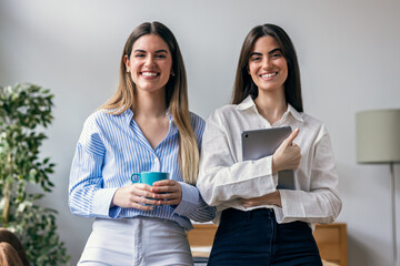 Two pretty business women taking a break while holding digital tablet looking at camera in the office