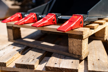 Heavy excavator bucket with sharp red steel teeth resting on a wooden pallet at a construction site, showcasing industrial machinery and digging equipment in detail