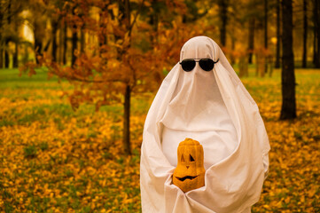 Woman in white sheet holding jack o'lantern against autumn forest background. Halloween costume. 