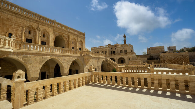 A panoramic view of ancient stone architecture in Mardin, Turkey, showcasing an intricate courtyard, arched windows, and traditional design under a bright, cloudy sky.