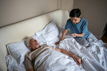 High angle view of worried older woman checking fever of sick husband in bed using thermometer....
