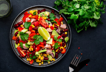 Tex-mex salad with canned tuna, avocado, corn, red beans, cherry tomatoes, cilantro and lime dressing, black table background, top view