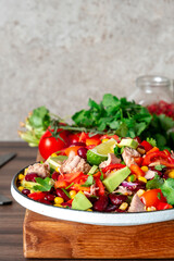 Preparing yummy salad with tuna, corn, avocado, beans, tomatoes and cilantro in plate on kitchen countertop and ingredients for preparation on wooden cutting board