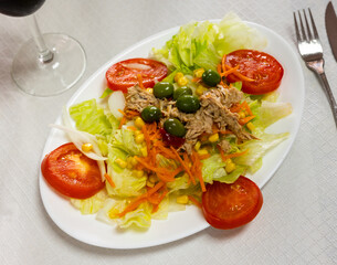 Appetizing salad of fresh greens, tomatoes, grated carrot, olives and canned tuna, served on white plate..