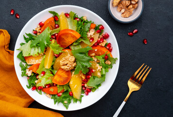 Autumn vegan persimmon salad with pomegranate seeds, walnuts, orange slices and lettuce, black background, top view