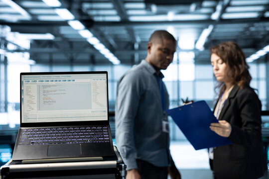 Lines of code on laptop screen in data center with programmers in blurry background. Close up of programming language on notebook display used by IT experts inspecting server clusters