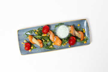 Red fish skewers with tomatoes, arugula, corn, and tartar sauce on a plate against a white background