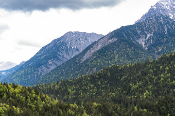 A dramatic alpine landscape with green hills and a sharp rocky mountain peak under a partly cloudy...