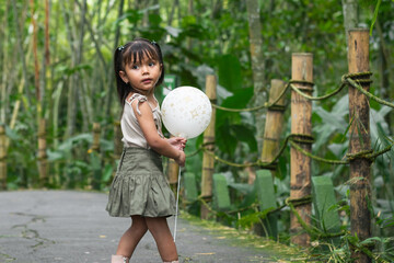 Latina toddler stands on zoo path holding white balloon and glances back amid lush bamboo forest.