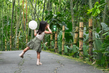 Latina toddler girl runs and look down zoo path clutching a white balloon in joyous stride amid bamboo forest