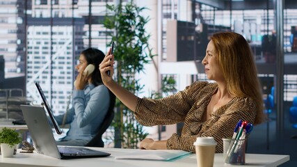 Confident general manager smiling and taking photos at her corporate workstation, using mobile...