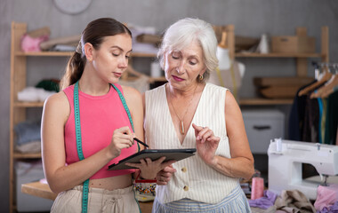 Elderly and adult seamstresses drawing pattern using tablet in sewing workshop