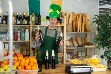 Cheerful salesman wearing green leprechaun hat and apron proudly offering bottles of beer,...