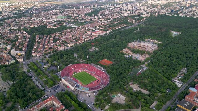 Aerial drone view of a large urban park hosting a packed football stadium, clay tennis courts, and a new stadium under construction, surrounded by dense green trees and city streets bustling with acti