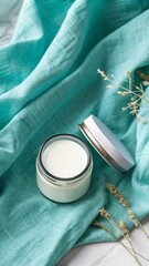 Cream container mockup on turquoise silk nylon fabric, light turquoise background with fabric and dried flowers.