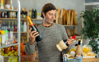 Casual middle-aged man thoughtfully comparing two wine bottles, examining labels and pondering choice with focused expression while shopping in grocery store