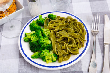 Green pasta with side of vibrant broccoli on white plate with blue trim, placed on checkered tablecloth. Healthy vegetarian dinner