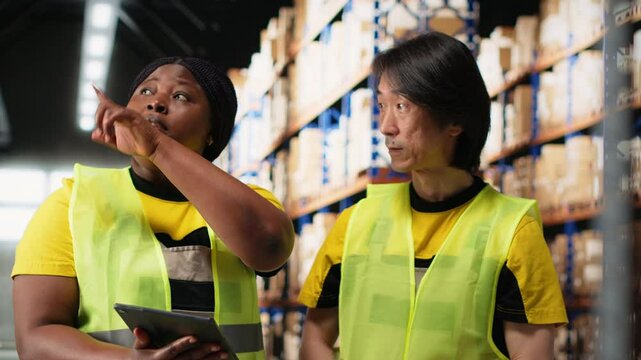 African american and asian team prepare shipments for dispatch with digital inventory and cargo pallets on depot racks. Working in a large scale distribution hub for express delivery. Camera B.