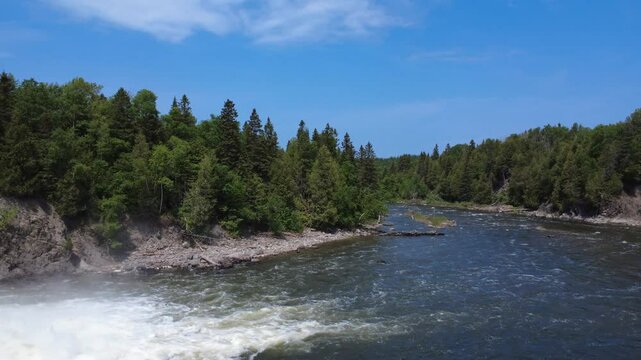 Drone footage soars above the Mitis River and lush green treetops under a clear blue sky in Price, Quebec, Canada. The river is renowned for sport salmon fishing.